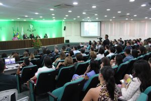 Vista do auditorio com pessoas assistindo palestra. Mesa de palestra tem 5 pessoas