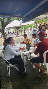 Defensor público Leandro Fulgêncio em serviço e expressão atenta, ouvindo demandas de moradores do bairro em uma mesa de plástico com computador