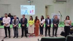 Quadro com defensores públicos em fileira ordenada diante a população que participa da audiência pública. No canto direito, a defensora Lia Felismino lê discurso para dar seguimento ao evento