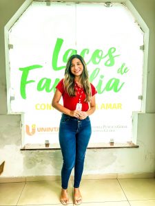 Na foto, ao centro, a estudante Eduarda Albuquerque posando sorridente para câmera. Atrás da mulher, está vitral com logomarca do programa Laços de Família